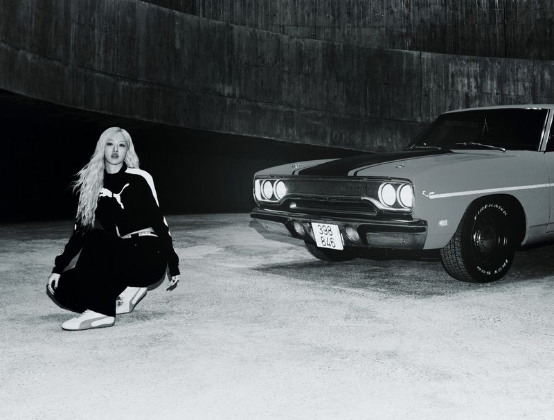 An adult female model in a Puma tracksuit is crouching in front of a vintage car with a dark urban background in a black and white photograph.