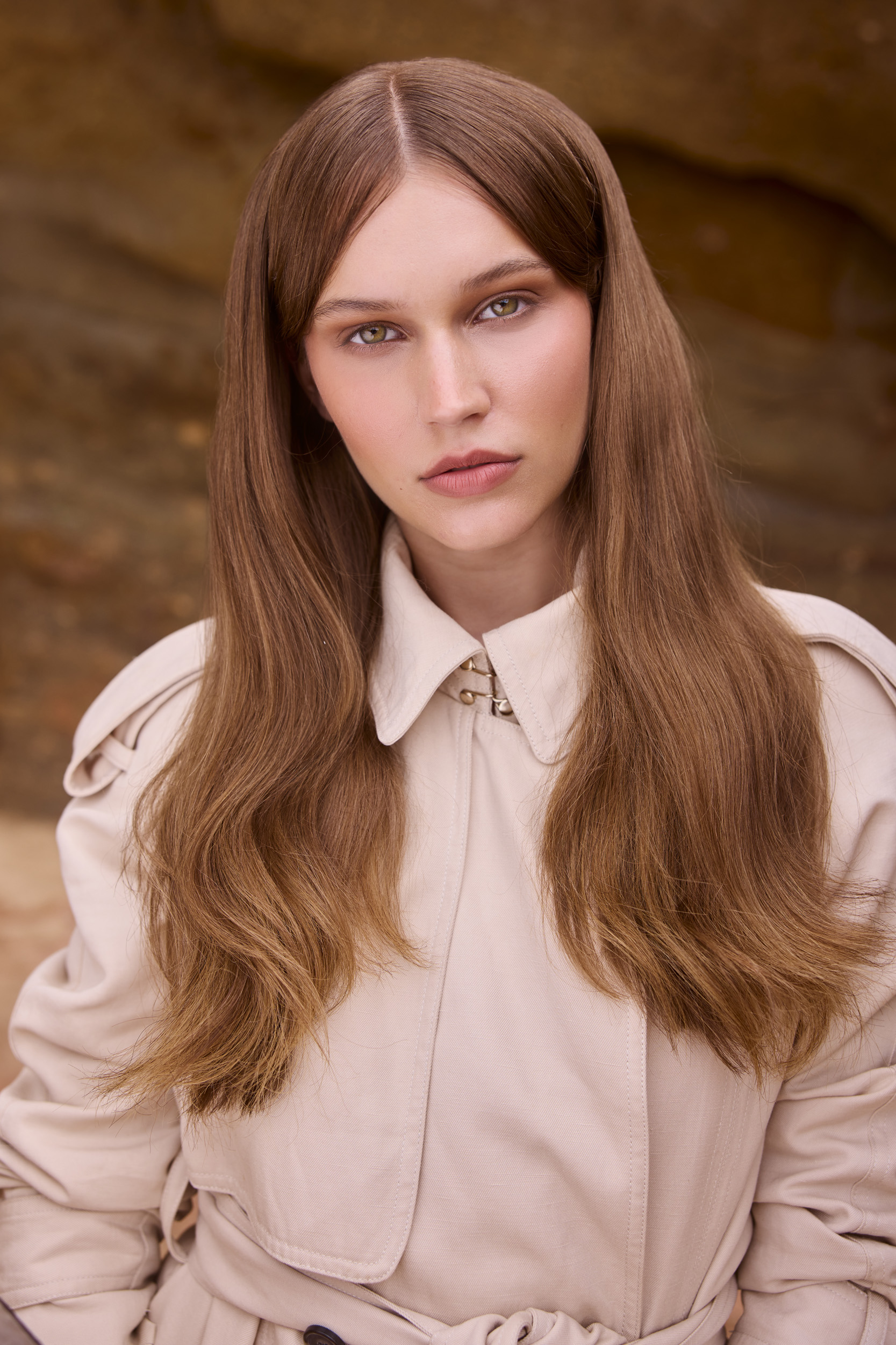 Portrait of a fair-skinned adult woman with long brown hair, wearing a beige trench coat, posing against a blurred brown background.