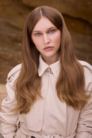 Portrait of a woman with long brown hair wearing a beige trench coat against a rocky background