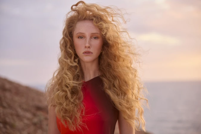 Close-up portrait of a woman with long blonde curly hair wearing a red top against an outdoor coastal background.