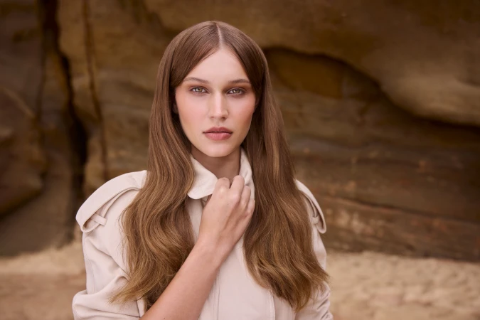 Portrait of a woman with long brown hair wearing a beige jacket against a rocky coastal background