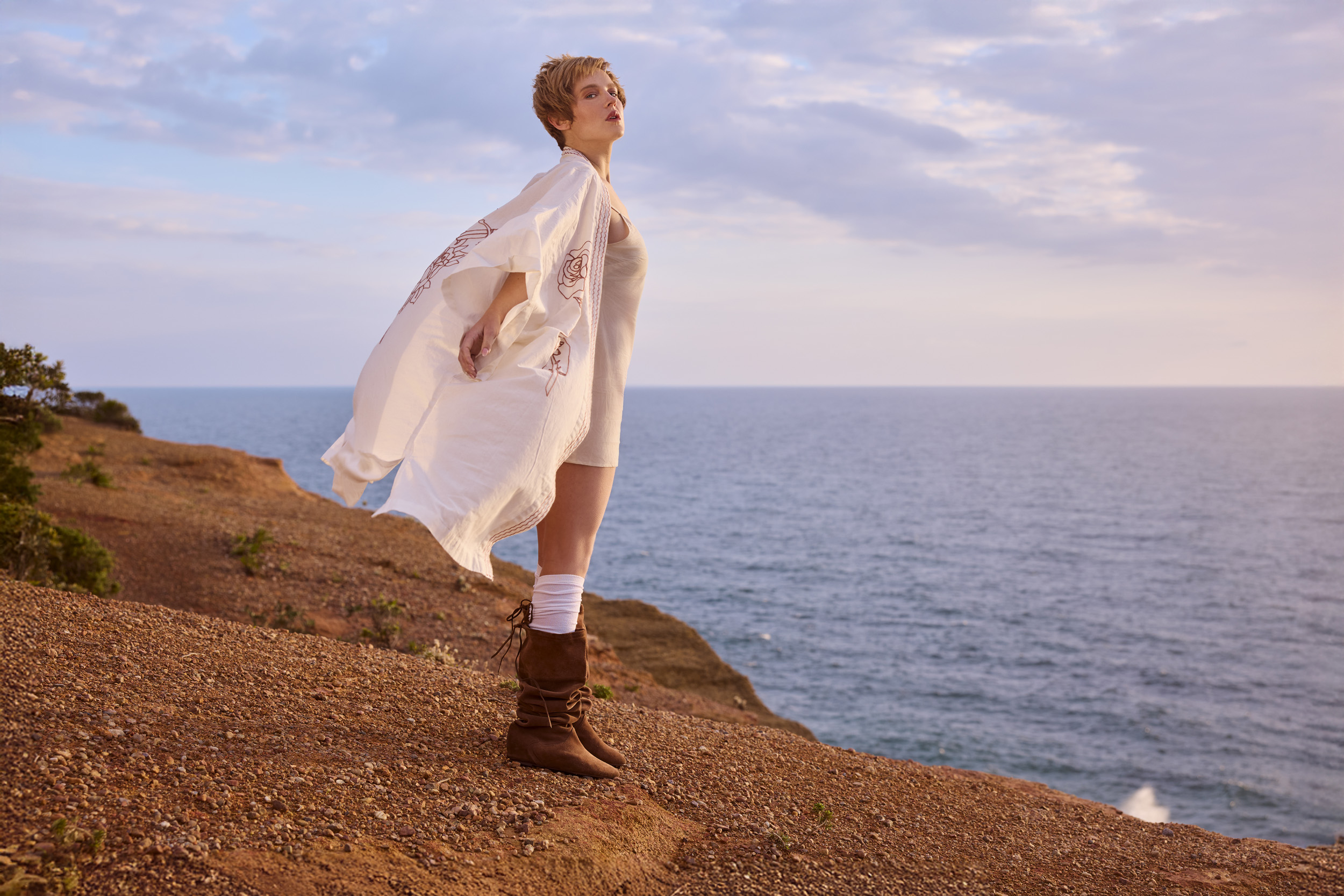A young adult female stands on a hillside overlooking the ocean at dusk, wearing boots and a white robe.