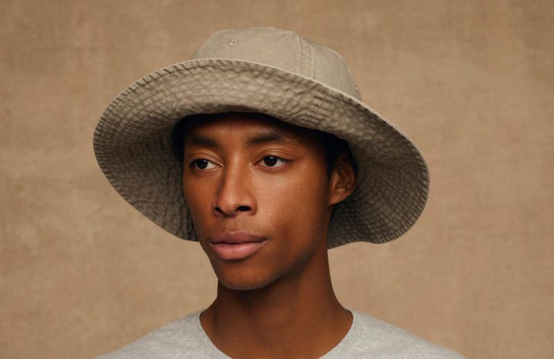 Close-up of a young adult male wearing a grey bucket hat and matching t-shirt against a neutral tan backdrop.