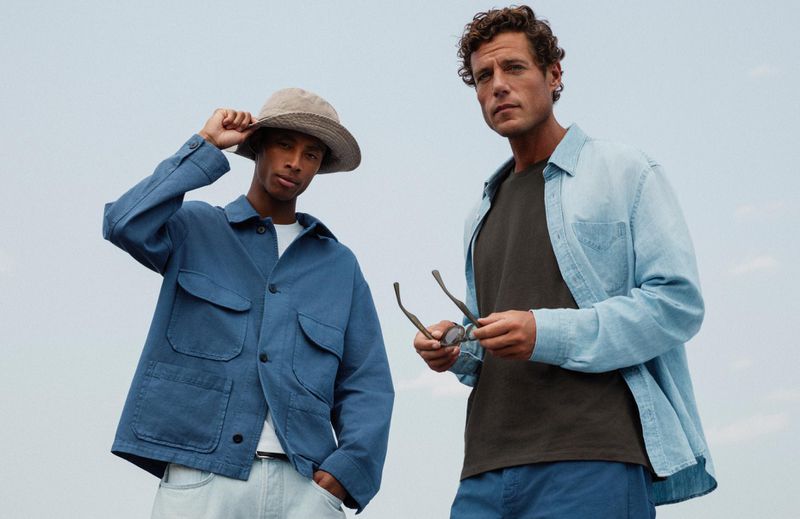Two adult males pose outdoors against a soft sky. One wears a blue chore coat and hat, the other a light blue shirt and holds glasses.