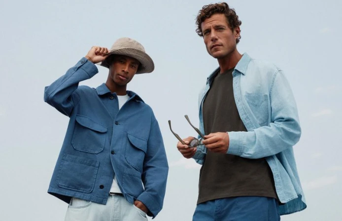 Two adult males pose outdoors against a soft sky. One wears a blue chore coat and hat, the other a light blue shirt and holds glasses.