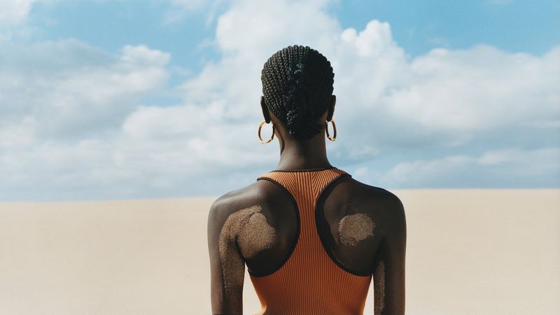 Back view of Black woman with braided hair in orange top standing on sandy beach with blue sky and white clouds, accessorized with gold hoops.