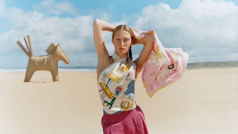 A fair-skinned woman in a pink skirt and patterned scarf poses on a beach with a wooden horse toy in the background.