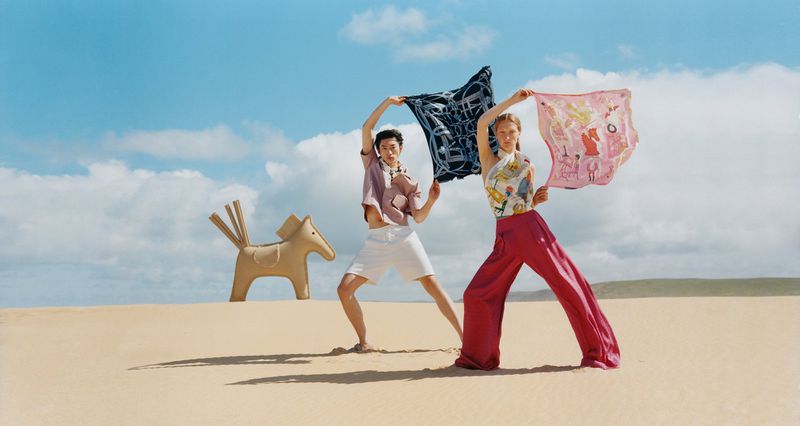 Two models in sand hold printed scarves, horse sculpture to left. Bright sky, sand dunes, colorful textiles, outdoor fashion shoot.