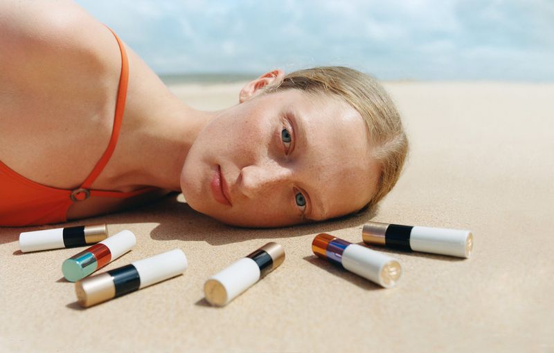 Woman in orange swimsuit lying on a sandy beach surrounded by cosmetic tubes. Beach scene with clear blue sky and neutral tones.