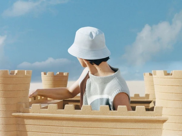 Person in bucket hat standing behind sandcastle against a blue sky. Fashion shot with neutral tones and seaside aesthetic.