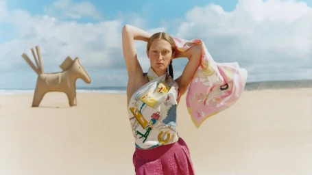 Model on a sandy beach holding a pink patterned silk scarf wearing a printed top and magenta skirt, with a large wooden horse sculpture behind.
