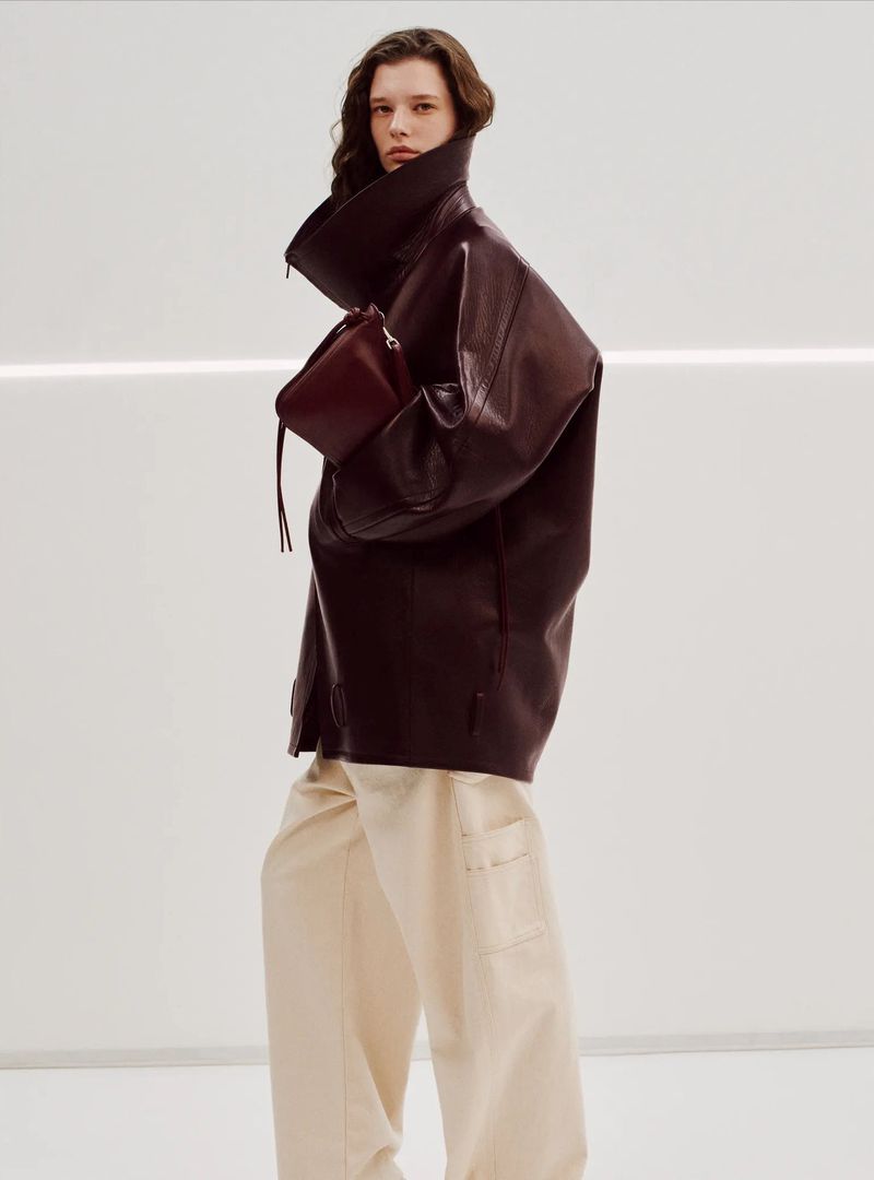 A woman in a dark leather coat, cream-colored pants, and holding a brown clutch poses in front of a white background with horizontal light.