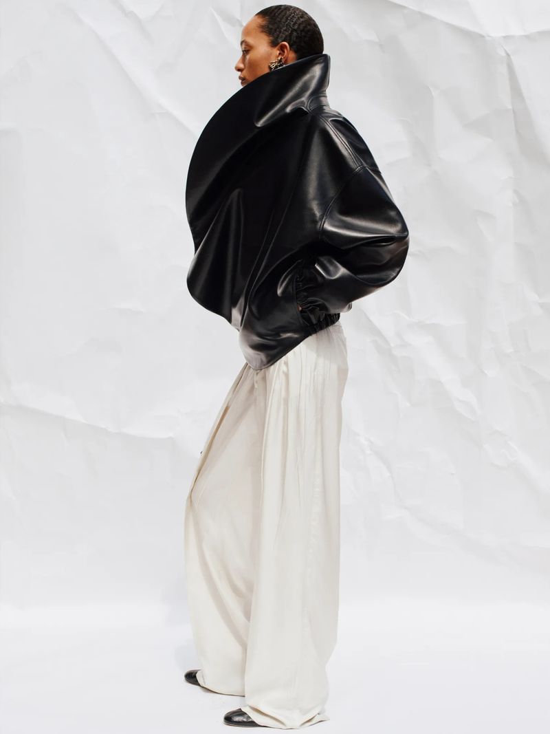 Fashionable woman poses in black leather jacket and cream wide-leg pants on white backdrop, side view.