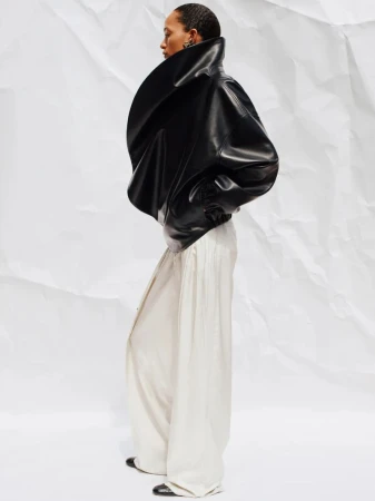 Fashionable woman poses in black leather jacket and cream wide-leg pants on white backdrop, side view.