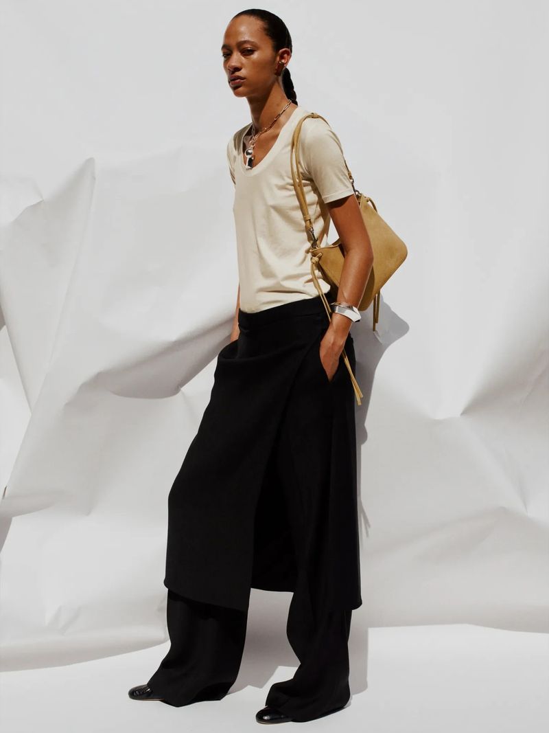 Fashion shoot featuring a woman wearing a beige t-shirt, black pants, jewelry and carrying a handbag, posing against a white background.