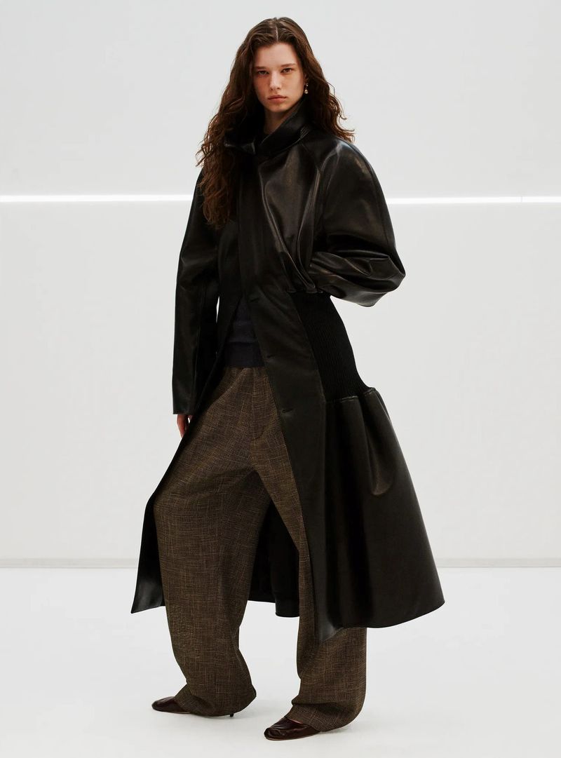Fashion model poses in studio wearing a black leather coat and brown checkered wide-leg pants against a white background with linear light.