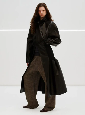 Fashion model poses in studio wearing a black leather coat and brown checkered wide-leg pants against a white background with linear light.