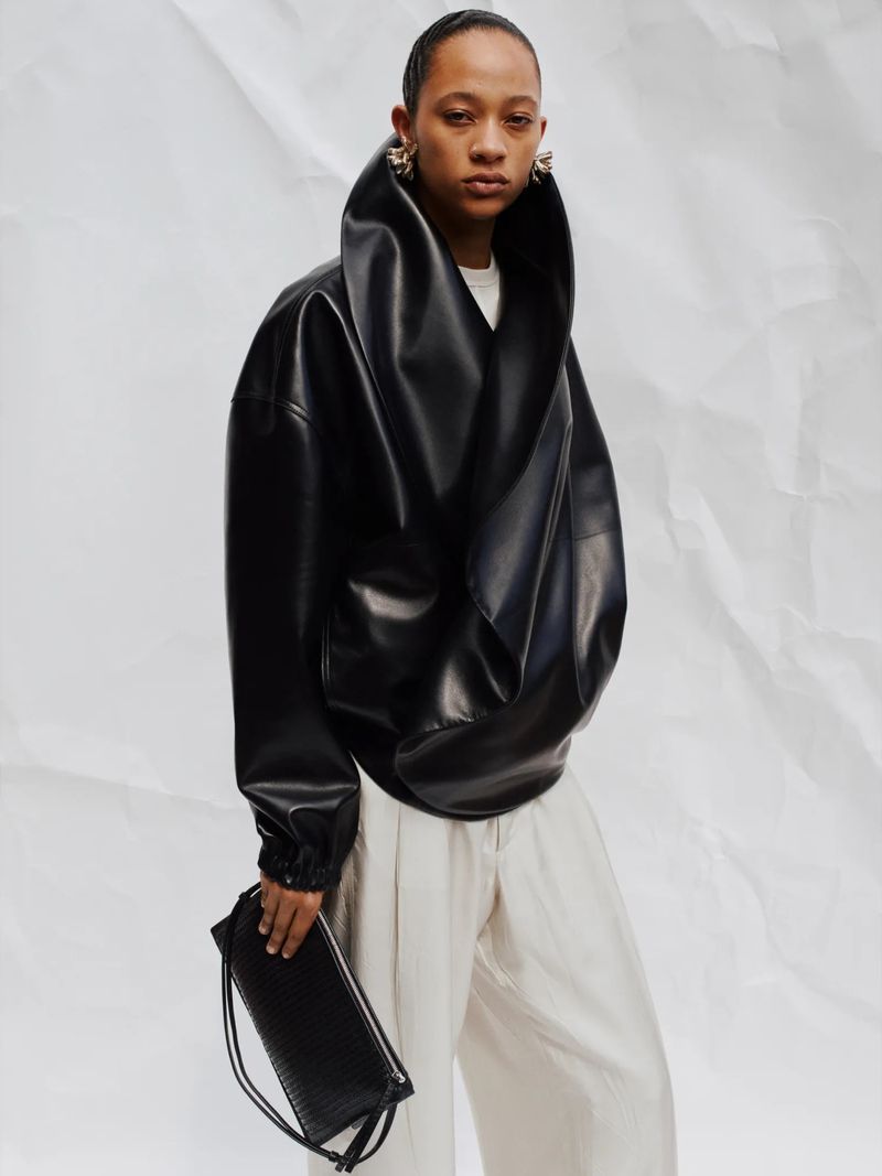 Adult woman poses in an oversized black jacket, light-colored pants, and dangling earrings, holding a black drawstring bag against a white background.