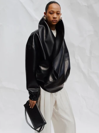 Adult woman poses in an oversized black jacket, light-colored pants, and dangling earrings, holding a black drawstring bag against a white background.