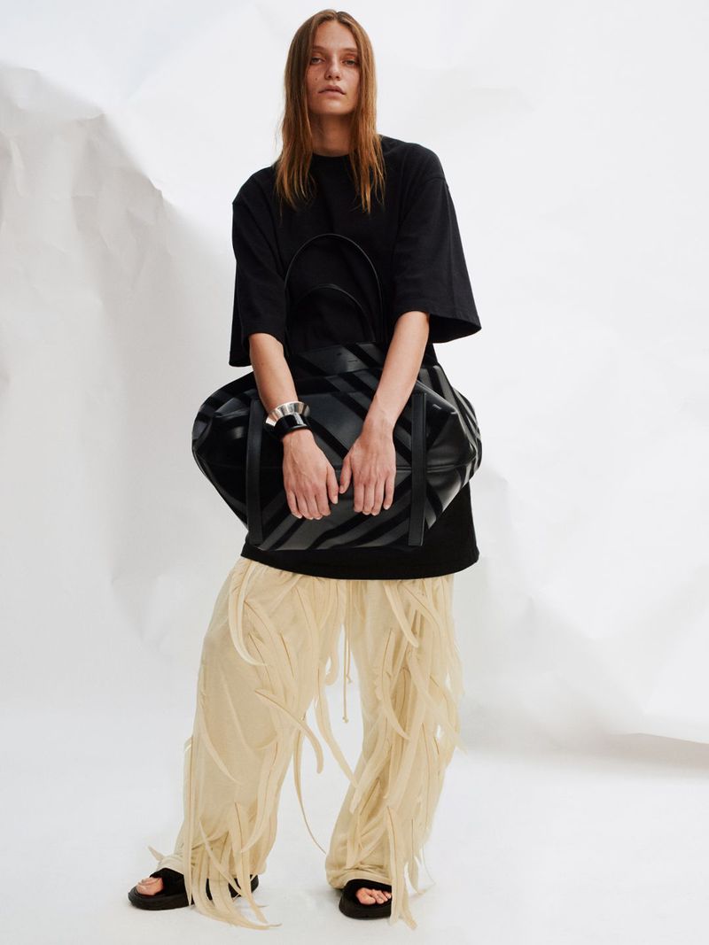 A fashion shot of a woman in a black T-shirt holding a striped tote bag, wearing beige pants with fringe, standing against a white background.