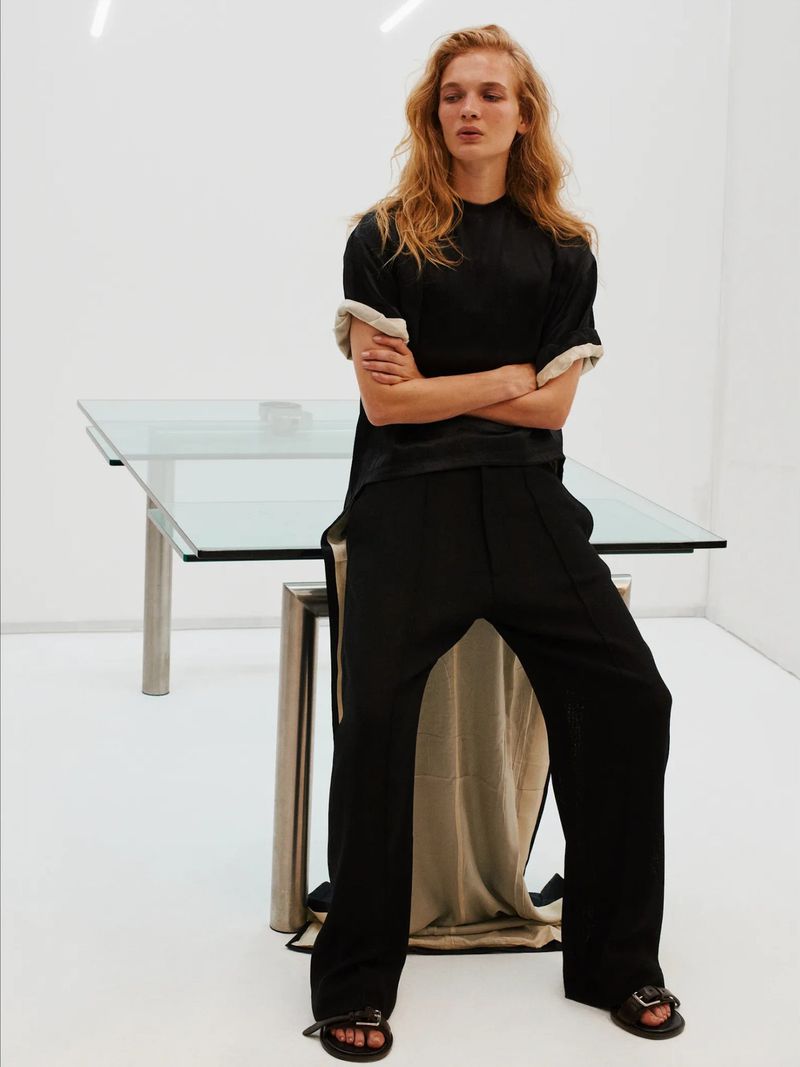 A fair-skinned female model poses in black t-shirt and trousers in a bright white studio with a glass-topped table beside her.