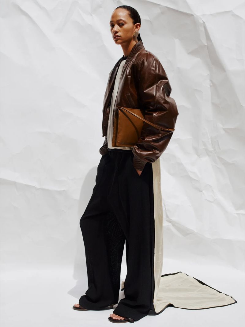 Studio shot of a fashion model wearing a brown leather jacket, light beige top, and black trousers against a white paper background.