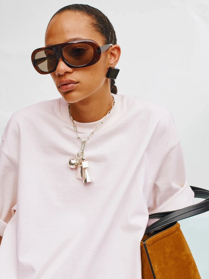 A woman poses wearing brown sunglasses and a silver chain necklace with charms, standing against a light backdrop, wearing a light pink T-shirt.