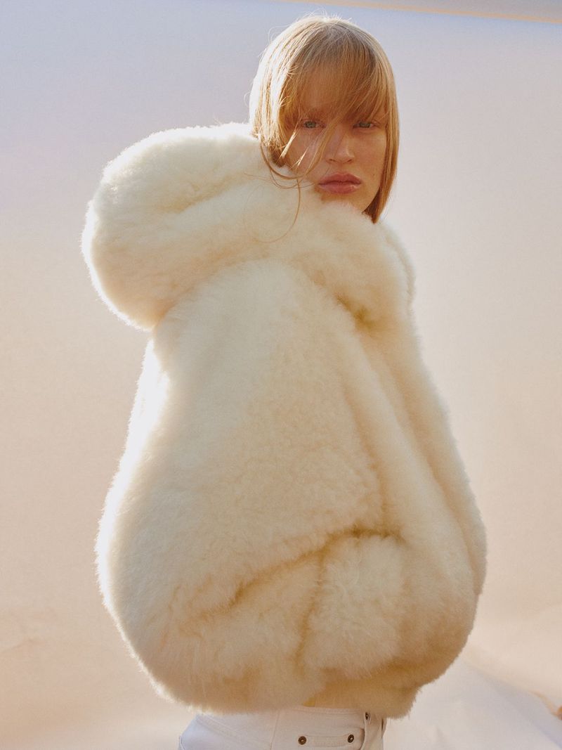 A woman poses in a fluffy hooded coat against a muted background, showcasing a fashion-forward look with neutral tones and soft textures.