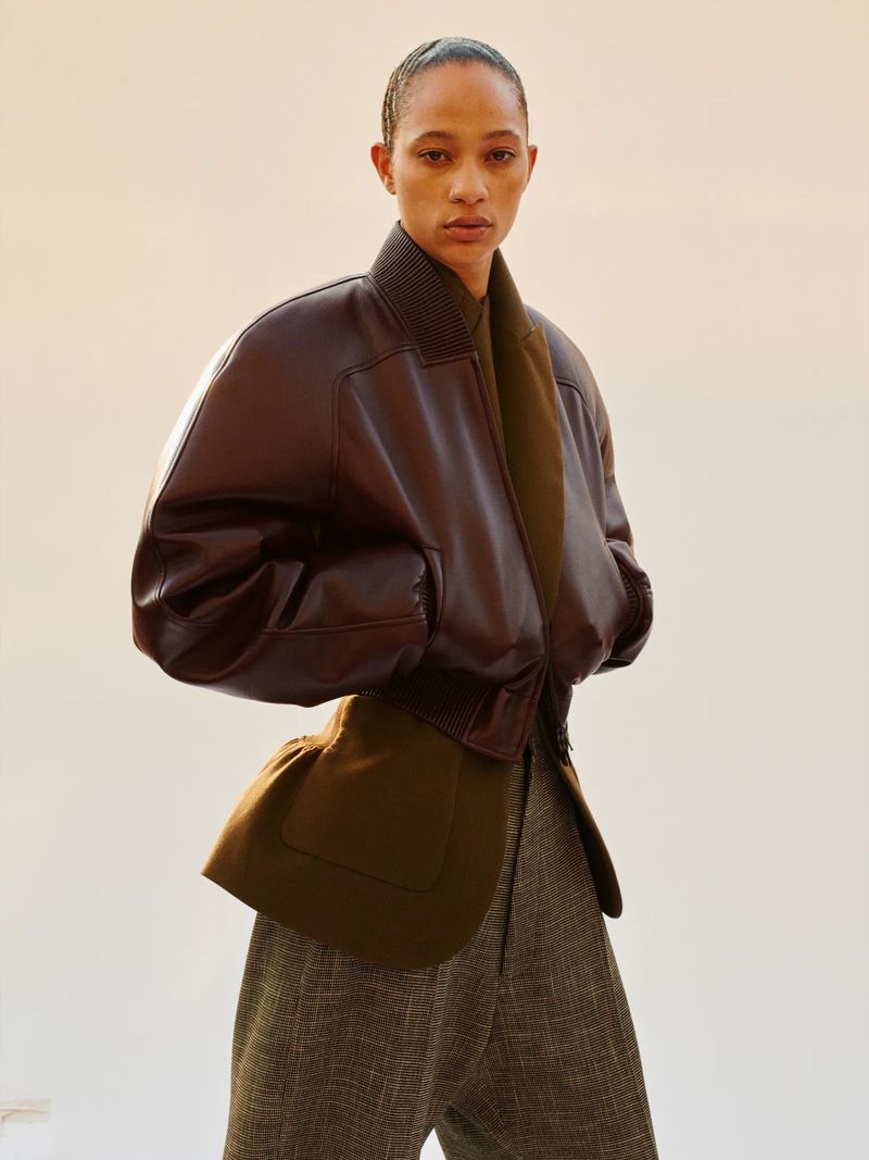 Fashionable adult woman poses in a studio wearing a brown leather jacket with green wool lining and tweed pants for a portrait.