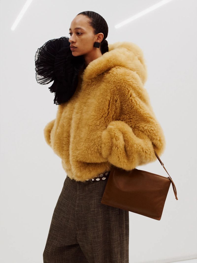 Woman in a hooded faux fur coat holding a brown clutch. She's wearing wide-legged plaid pants in a minimalist indoor setting.