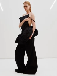 Model wearing dark sunglasses, black structured top, and wide trousers standing against a plain white studio backdrop.