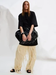 Model in black t-shirt and cream fringed trousers holding a large black leather bag against a white seamless backdrop.