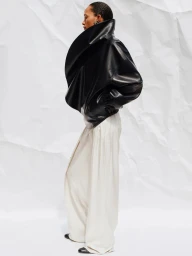 Model wearing a structured black leather jacket and white wide-leg trousers against a crinkled white backdrop