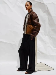 Model in brown leather jacket and black trousers holding a tan leather clutch against a rumpled white paper backdrop.
