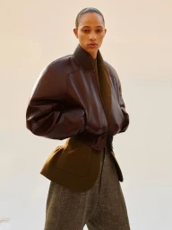 Model wearing a dark brown leather bomber jacket and grey trousers against a clean, neutral background.