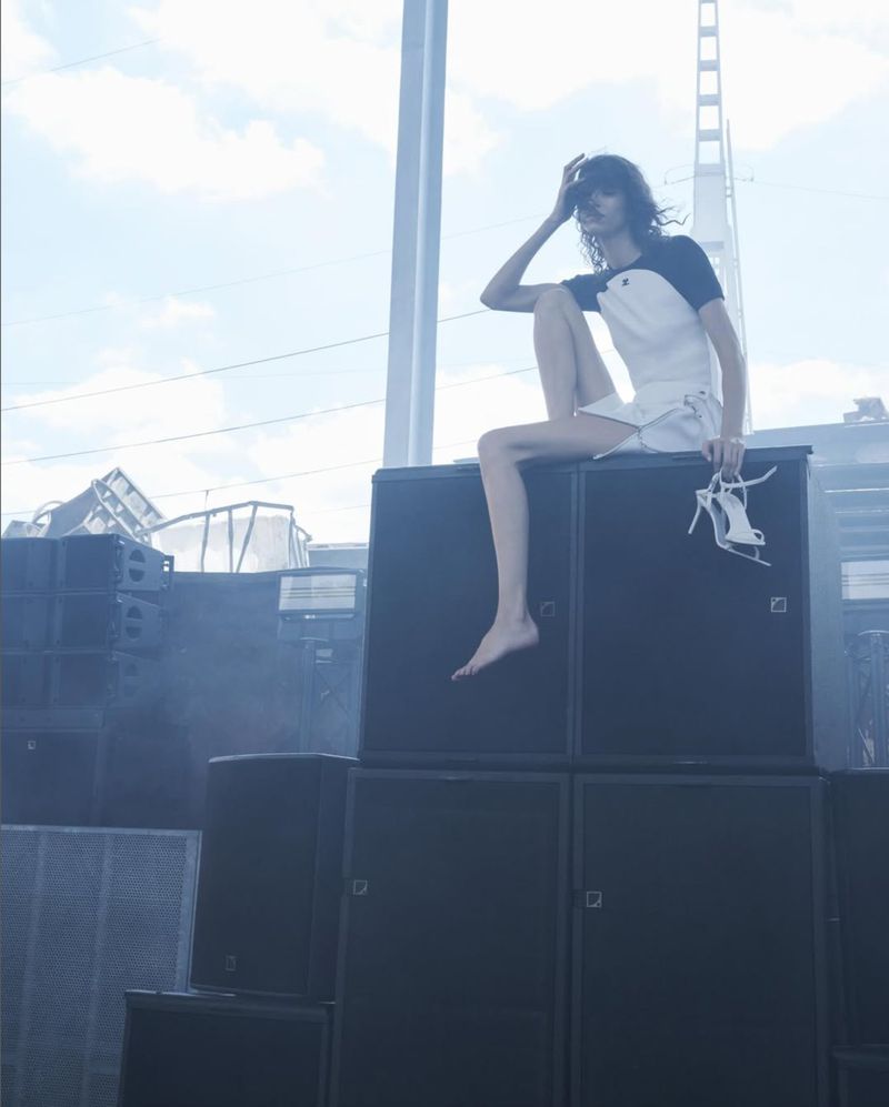 A bare-footed woman with white shorts and a top sits on a stack of black speakers, holding white high heels, under a blue sky.