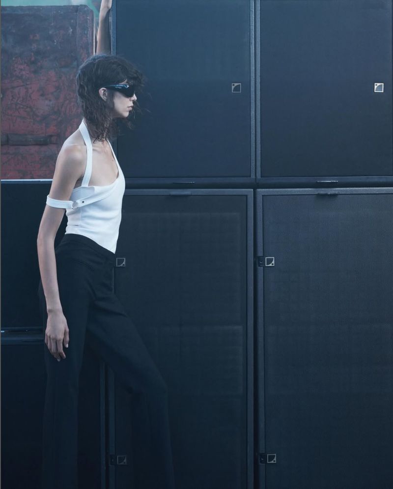A side-angle shot of a woman in a white halter top and dark pants posing near a wall of dark, stacked speakers.