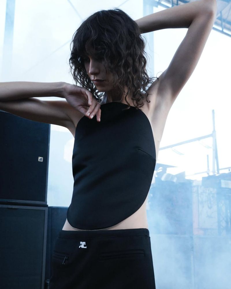 A woman poses in a black top and pants with her arms above her head in front of a blurred industrial background with speakers.