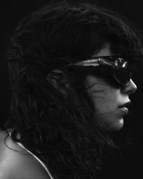 Model wearing dark sunglasses and dark clothing against a black background in a profile portrait