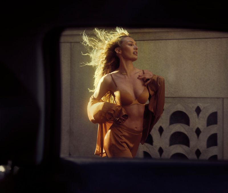 A blonde female model in a gold two-piece outfit and jacket poses in front of a stone wall, framed by the edge of a dark car window.