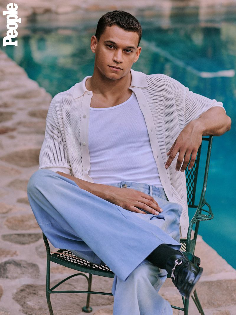 A young adult male sits on a chair outdoors, wearing a white top, open sweater, light blue pants, and black shoes, posing for a portrait.