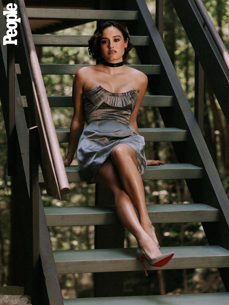 Portrait of a woman in a strapless dress sitting on wooden stairs outside, styled with a choker and red-bottom heels in natural light.