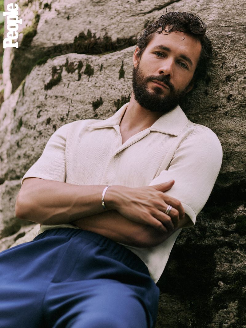 Portrait of an adult man with a beard, wearing casual clothing, leaning against a rock, and looking at the camera.