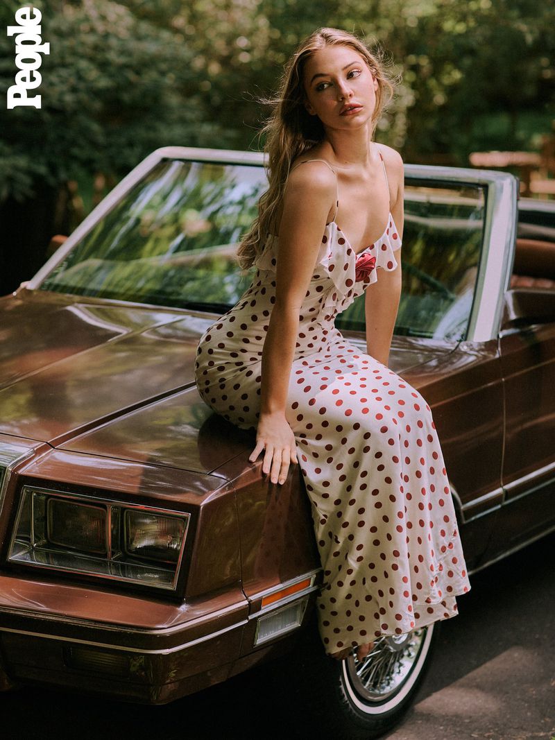 A blonde woman in a polka dot dress sits on the hood of a brown convertible car surrounded by green trees and foliage.