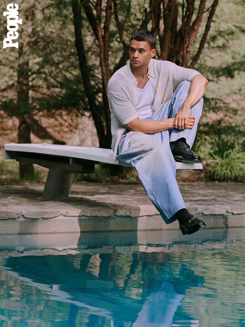 A man sits on a diving board above a pool. He wears a light shirt, loose blue pants, and loafers. The background includes trees and sky.