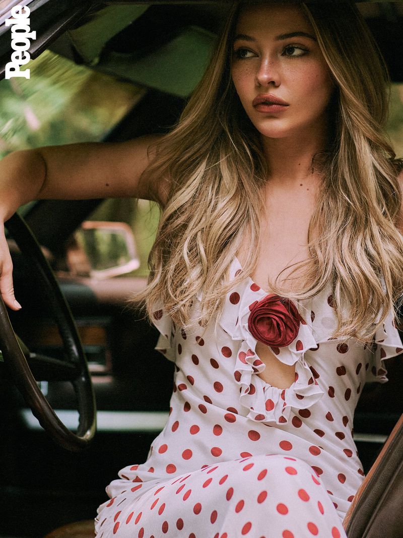 Indoor portrait of a young woman with long blonde hair, wearing a white dress with red polka dots, seated in a vintage car.