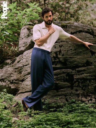 Portrait of adult male posing in nature, wearing a light-colored shirt and dark blue pants next to a rock in a natural, wooded setting.