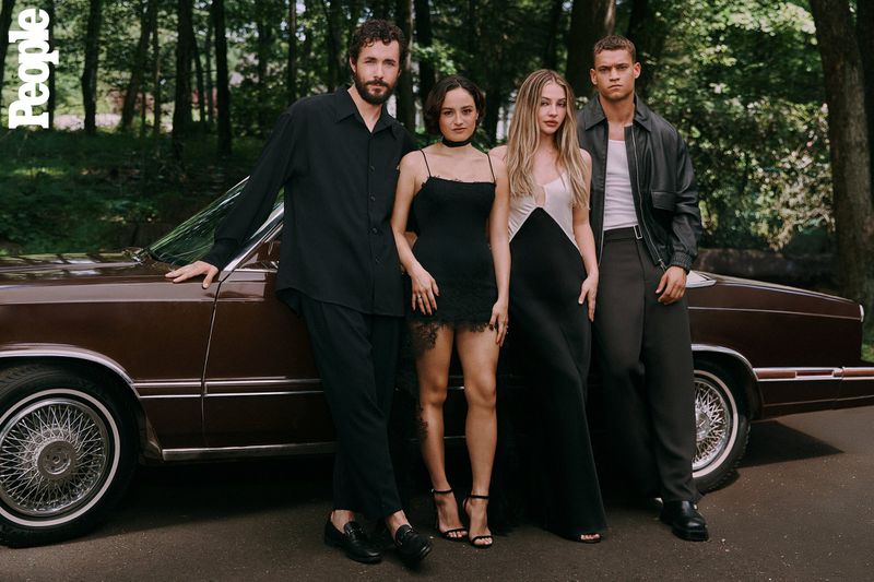Four young adults pose with a vintage brown car in a wooded area. Two women wear dresses, while the two men sport stylish monochrome outfits.