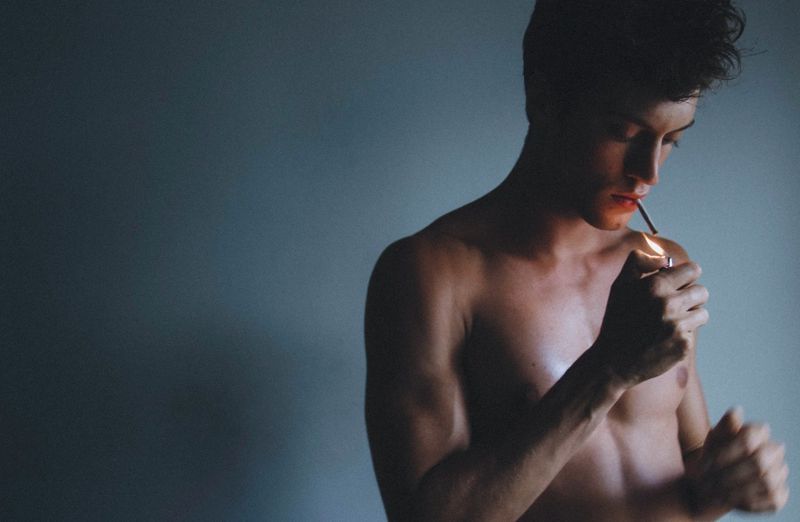 Portrait of shirtless adult male lighting a cigarette in a dark indoor room with soft lighting and a simple gray background.