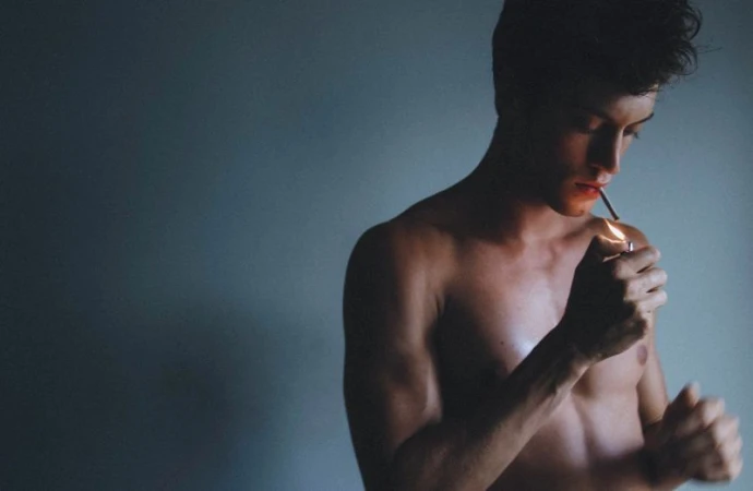 Portrait of shirtless adult male lighting a cigarette in a dark indoor room with soft lighting and a simple gray background.